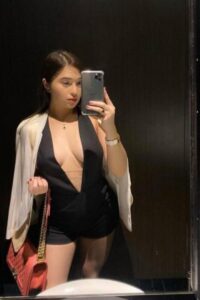 KL Escort Girl Woman in a black romper taking a mirror selfie with a pink purse and white jacket. Malaysia Sex Service