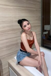 KL Escort Girl Woman in denim shorts and burnt orange top sitting on bathtub edge. Malaysia Sex Service